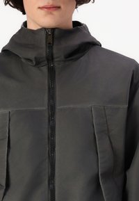 Mørk grå hettejakke med frontglidelås og to vertikale lommer. Materialet ser glatt og noe glinsende ut med en tettsittende design.