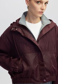 Chaqueta marrón con una sección superior texturizada, cuello gris, cintura con cordón y cierre frontal. Tiene bolsillos laterales y puños elasticados.
