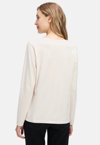 Bluzka z długim rękawem z jasnobeżowej tkaniny. Charakteryzuje się szerokim dekoltem i miękką fakturą, prostym krojem oraz minimalistycznymi detalami.