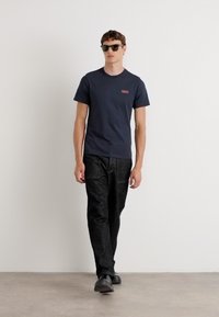 Tricou din bumbac de culoare navală cu un mic logo roșu, asortat cu pantaloni texturați negri și pantofi slip-on negri, purtat de un model masculin.