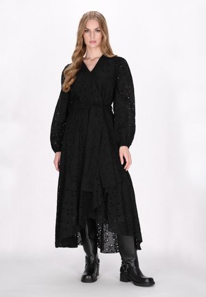 Schwarzes Wickelkleid mit langen Puffärmeln, aus strukturiertem Stoff mit verstreuten Löchern. Kombiniert mit kniehohen schwarzen Lederstiefeln.