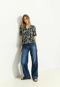 Sort kortærmet bluse med hvide paisley-mønstre, parret med løse blå denimjeans og sorte sandaler på en lys baggrund.