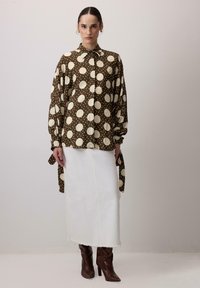 Blusa marrone a pois con chiusura a bottoni e maniche oversize, abbinata a una gonna di jeans bianca e stivali alti marroni.