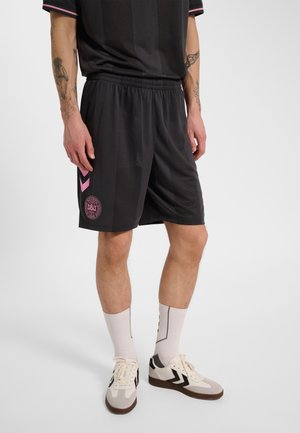 Mężczyzna ubrany w czarne sportowe spodenki z różowym logo i akcentami w kształcie wzoru jodełki, białe skarpety i białe buty sportowe z czarnymi detalami.