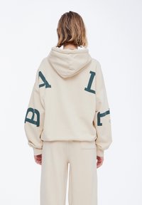 Hoodie beige de gran tamaño con capucha ajustable y letras gráficas en color verde oscuro en las mangas y la espalda. Tejido suave, ajuste relajado y mangas largas.