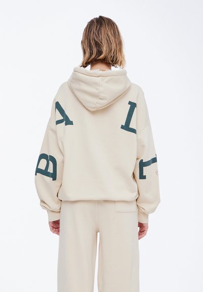 Beige oversized hettegenser med snøring og mørkegrønne grafiske bokstaver på ermene og ryggen. Mykt stoff, avslappet passform og lange ermer.
