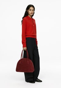 Roter Wollpullover mit Kragen und Knöpfen, kombiniert mit schwarzer Hose im weiten Schnitt. Trägt eine burgunderfarbene Wildleder-Handtasche mit runder Form und doppelten Griffen.