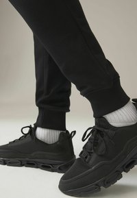 Las zapatillas deportivas negras cuentan con una parte superior de malla y sintético, suela acolchada y cordones redondeados. Combinadas con joggers negros ajustados y calcetines acanalados.