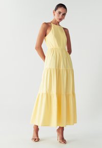 Nainen, jolla on ylläänolkaimeton, kerroksellinen, vaaleankeltainen maksimekko, jossa on kaula-aukko ja beige avoin kärki -korkokenkä, seisoo yksinkertaista valkoista taustaa vasten.