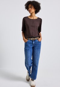 Bruine suedekorte mouwen top, blauwe wijde jeans, een luipaardprintriem en witte sneakers met bruine accenten. Eenvoudige, casual outfit.