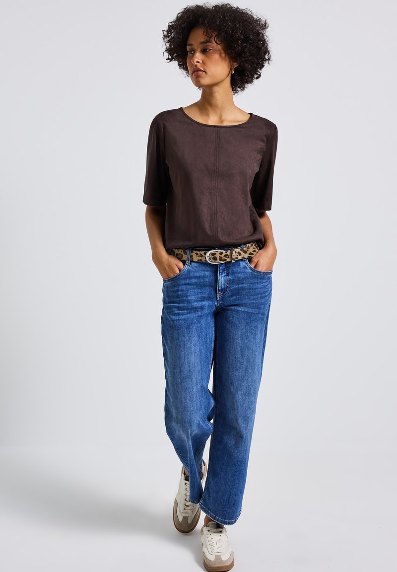 Bruine suedekorte mouwen top, blauwe wijde jeans, een luipaardprintriem en witte sneakers met bruine accenten. Eenvoudige, casual outfit.