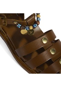 Braune Ledersandale mit breiten Riemen, goldfarbener Hardware und dekorativen Perlenakzenten mit blauen und natürlichen Holzperlen.