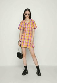 Vestido xadrez de manga curta em rosa e amarelo com fecho de botões, combinado com botas de combate pretas e uma pequena bolsa preta.