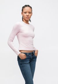 Jonge persoon met gevlochten haar, draagt een lichtroze aansluitende lange mouwen top en een blauwe spijkerbroek, staand met handen in de zakken.