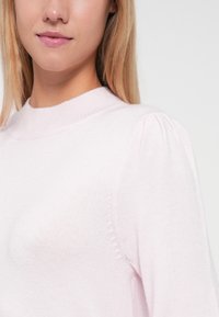 Gaiši rozā džemperis ar augstu kaklu, mīkstu tekstūru un pūkainām piedurknēm. Satur smalkas šuvju detaļas gar šuvēm.