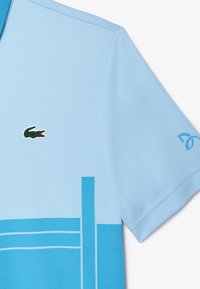 Pikétröja i ljusblått med mörkblå ränder. Har en grön krokodil-logotyp på bröstet och unika detaljer på ärmarna.