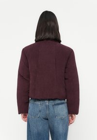Persoon met steil, donker haar die een kort, donkerpaarse corduroy jack en hoge blauwe spijkerbroek draagt, met de rug naar ons gekeerd.