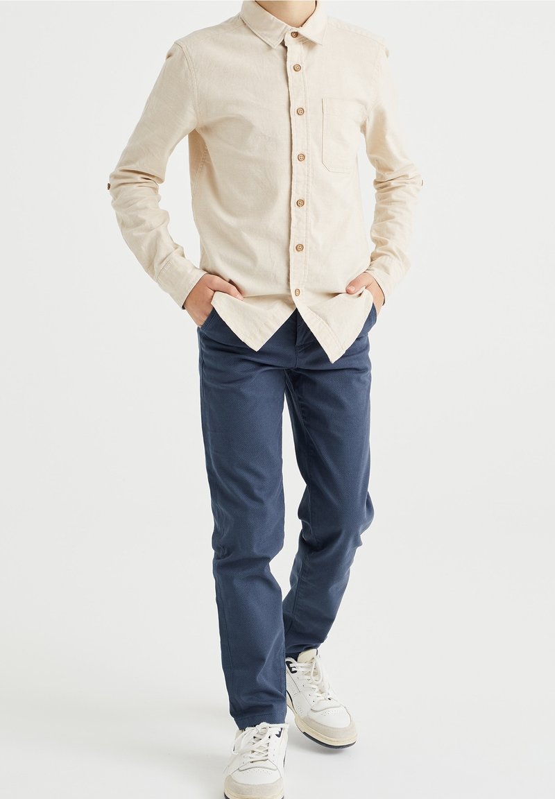 Světle béžová košile na knoflíky s přední kapsou, spojená s tmavě modrými, slim-fit kalhotami. Bílé tenisky doplňují celý outfit.