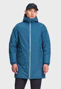Homme portant une longue veste d'hiver bleue à capuche avec une fermeture éclair à l'avant et une casquette noire, debout devant un fond uni.