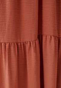 Rostfärgad tyg med mjuk textur, med samlade sömmar som skapar en volangnederkant. Slät, lätt material.