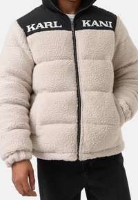 Giacca puffer in pile beige con spalle in nylon nero. Presenta una zip frontale, colletto alto e il testo del logo "KARL KANI" sul petto.