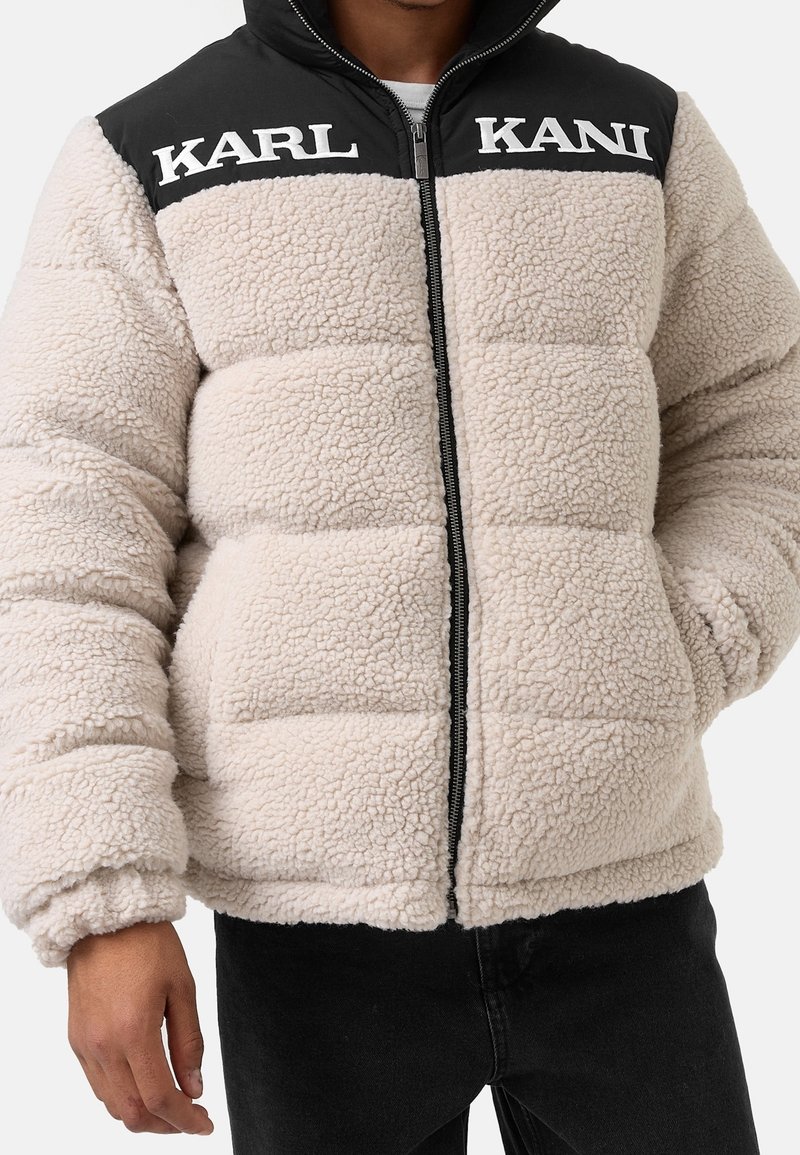 Giacca puffer in pile beige con spalle in nylon nero. Presenta una zip frontale, colletto alto e il testo del logo "KARL KANI" sul petto.