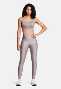 Ljus mauve sportbh och högmidjade leggings, tillverkade av mjukt, töjbart material med logotypdetaljer. Modellen står med händerna på höfterna.