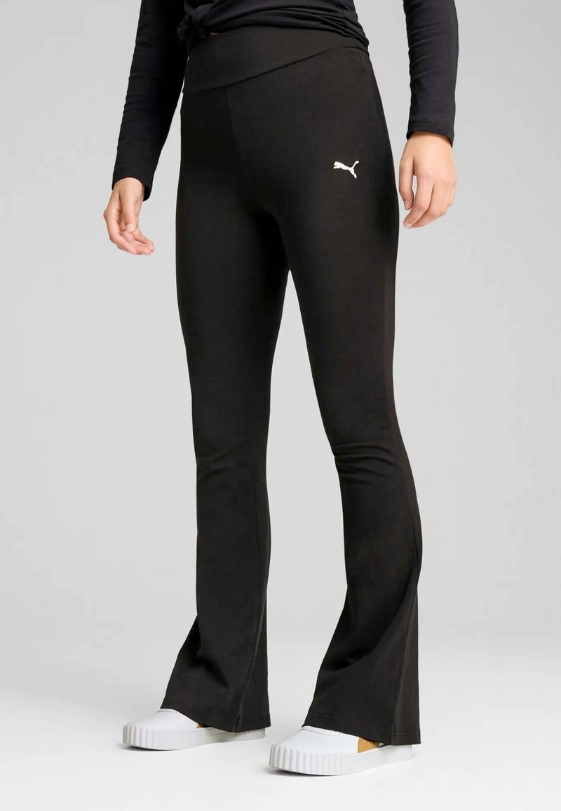 Persoon in zwarte Puma flared leggings en witte instappers, staand tegen een effen grijze achtergrond.