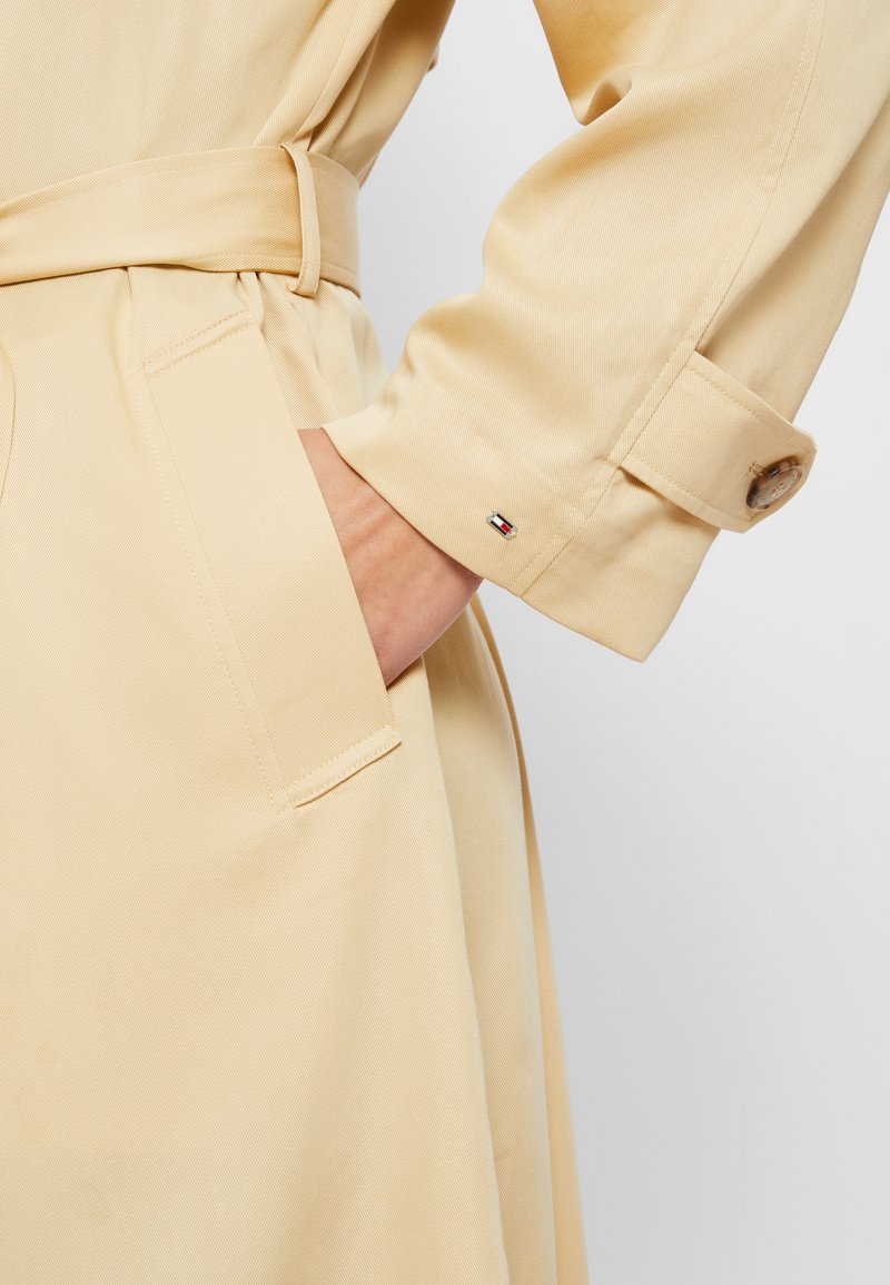 Trench coat beige con vita evidenziata da una cintura, dotato di una tasca laterale e polsini con bottoni. Un logo in evidenza è visibile vicino al polsino. Tessuto con una texture liscia.