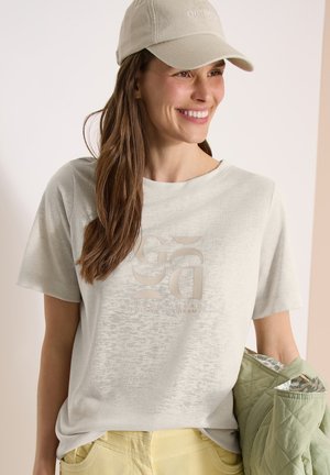 Femme souriante portant une casquette beige et un t-shirt beige clair avec un texte subtil, tenant une veste matelassée verte au niveau de la taille.
