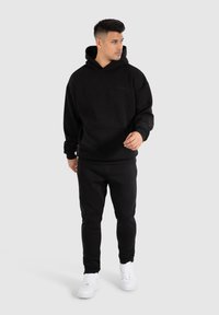 Un sweat à capuche noir et un pantalon de survêtement assorti en matériau doux. Il présente une poche avant, une coupe décontractée et un design minimaliste. Accompagné de baskets blanches.