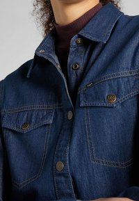 Dunkelblaue Jeanshemd mit Button-Down-Kragen, zwei Fronttaschen und gebrandeten Knöpfen. Verfügt über sichtbare Nähte und eine weiche Textur.