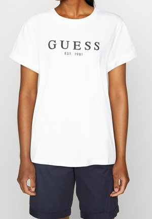 Persoon die een wit T-shirt draagt met de zwarte tekst "GUESS EST. 1981" en donkerblauwe kniebroek tegen een effen achtergrond.