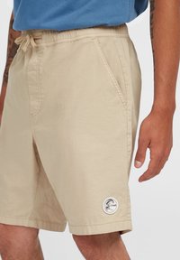 Beige casual shorts with drawstring waist and front pockets worn by a person with tattooed arms and a blue shirt.