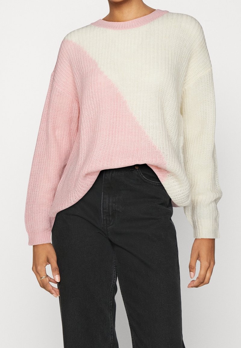 Pull en tricot arborant un motif diagonal rose et blanc, col rond et manches longues avec poignets côtelés. Porté avec un jean foncé.