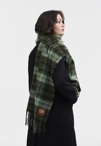 Sciarpa a scacchi verdi realizzata in materiale morbido, con motivi a contrasto nero e verde, bordi frangiati e una piccola etichetta in pelle marrone.