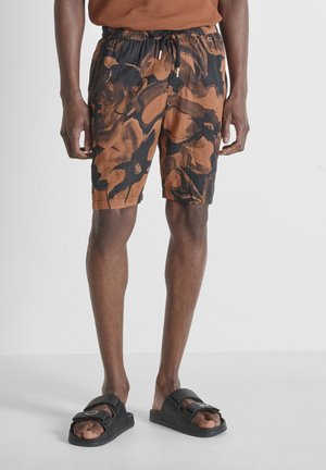 Uomo che indossa pantaloncini con stampa astratta marrone e nera e sandali neri con fibbia, in piedi davanti a uno sfondo grigio chiaro uniforme.