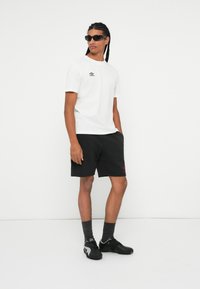 Moški z narezanimi lasmi in sončnimi očali, oblečen v belo majico Umbro, črne kratke hlače, temne nogavice in črne superge Puma, stoji ob beli steni.