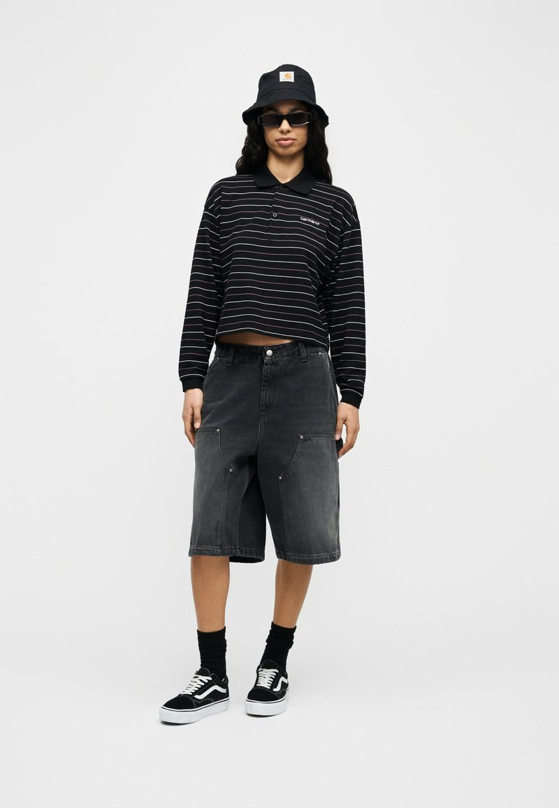 Persoon met zwarte vissershoed, rechthoekige zonnebril, zwarte gestreepte cropped longsleeve, donkere knielange korte broek en zwarte sneakers.