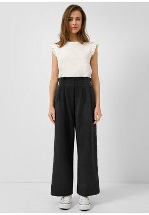 Kobieta stojąca w białym topie bez rękawów z koronkowym wykończeniem, czarnych spodniach o wysokim stanie i szerokich nogawkach oraz białych trampkach na jednolitym szarym tle.