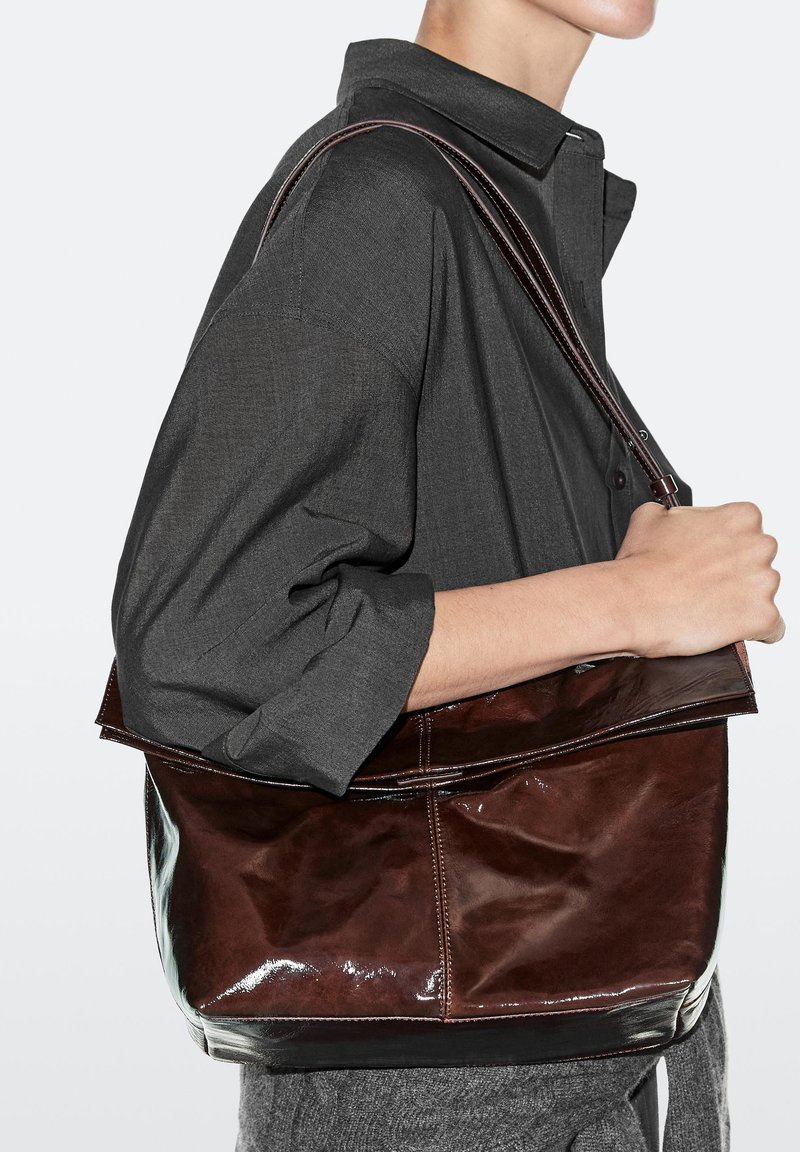 Person trägt dunkelgraues, geknopftes Hemd und graue Hose, trägt eine große glänzende dunkelbraune Leder-Schultertasche.