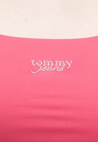 Tecido canelado rosa vibrante com um logótipo "tommy jeans" bordado a prata no centro. Vista em close-up que destaca a textura e os detalhes.
