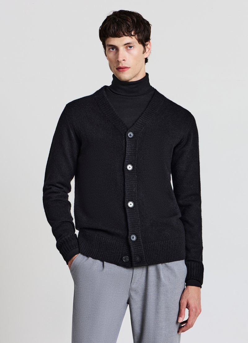 Schwarze Strickjacke mit V-Ausschnitt, Knopfverschluss und gerippten Bündchen und Saum. Getragen über einem schwarzen Rollkragenpullover, kombiniert mit hellgrauen Hosen.