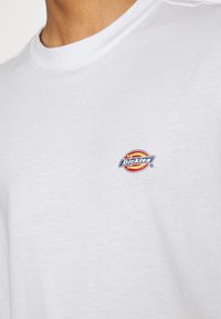 Bijela pamučna majica s malim, višebojnim „Dickies“ logotipom na lijevoj strani prsnog područja. Ima okrugli izrez i glatku teksturu.