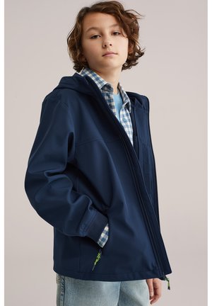 Junge Person mit kurzen braunen Haaren trägt eine marineblaue Kapuzenjacke über einem blau karierten Hemd und hellblauen Jeans, steht vor einfachem Hintergrund.