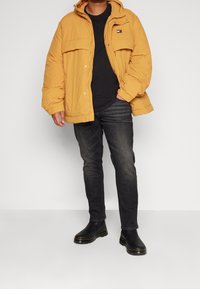 Gul vadderad jacka med huva, två framfickor och logotypdetalj. Svart t-shirt under och mörka jeans. Svarta ankelstövlar.