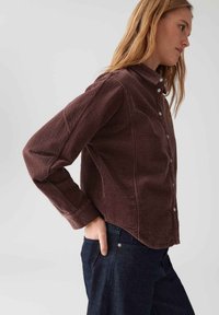 Vrouw in een donkerbruine corduroy button-up shirt met lange mouwen, zijwaarts staand met één hand in de zak van haar donkerblauwe spijkerbroek tegen een effen achtergrond.