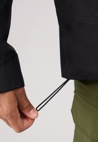 Cuff di una giacca nera con un cordoncino tenuto da una mano. Il tessuto sembra liscio con un design strutturato e una finitura opaca sottile.