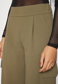 Pantaloni tailored de culoare verde măsliniu, cu talie înaltă și pliseuri; au buzunare laterale și un finisaj de țesătură texturată, netedă.