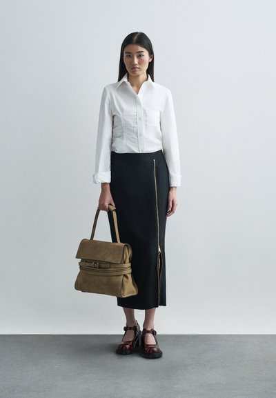 Witte blouse met gestructureerde stof, zwarte rok met een zijrits, tan tas en bordeauxrode schoenen met een bandje, op een grijze vloer.